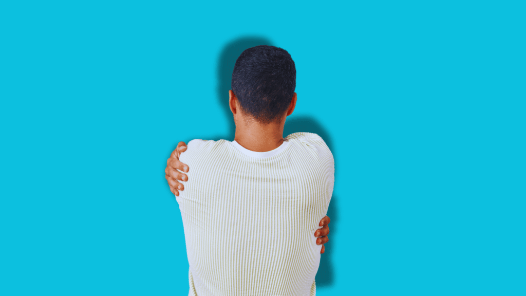 Person seen from behind wearing a white ribbed sweater, arms wrapped around themselves in a self-hug against a bright turquoise blue wall, representing self-comfort and the need for self-compassion
