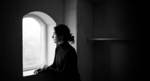 A high-contrast black and white photograph of a woman in a turtleneck, viewed in profile. She is standing at a deep, arched window and gazing thoughtfully outside into a bright, overcast light, framed by shadow. Her expression is calm and introspective.